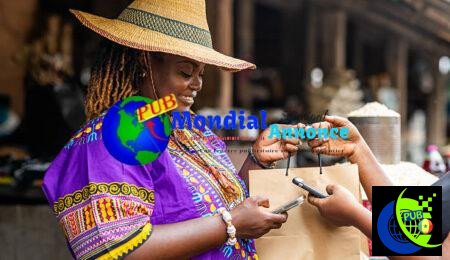 Small business owner, SME. Local traditional African market woman in a purple dashiki and straw hat receives contactless mobile money phone payment transactions at her vibrant outdoor market stall. Téléphone, Internet et petites annonces : le trio gagnant du commerce local